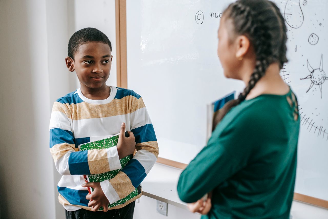 Smart African American schoolboy standing near whiteboard with stack of textbook and answering question with anonymous ethnic classmate during lesson