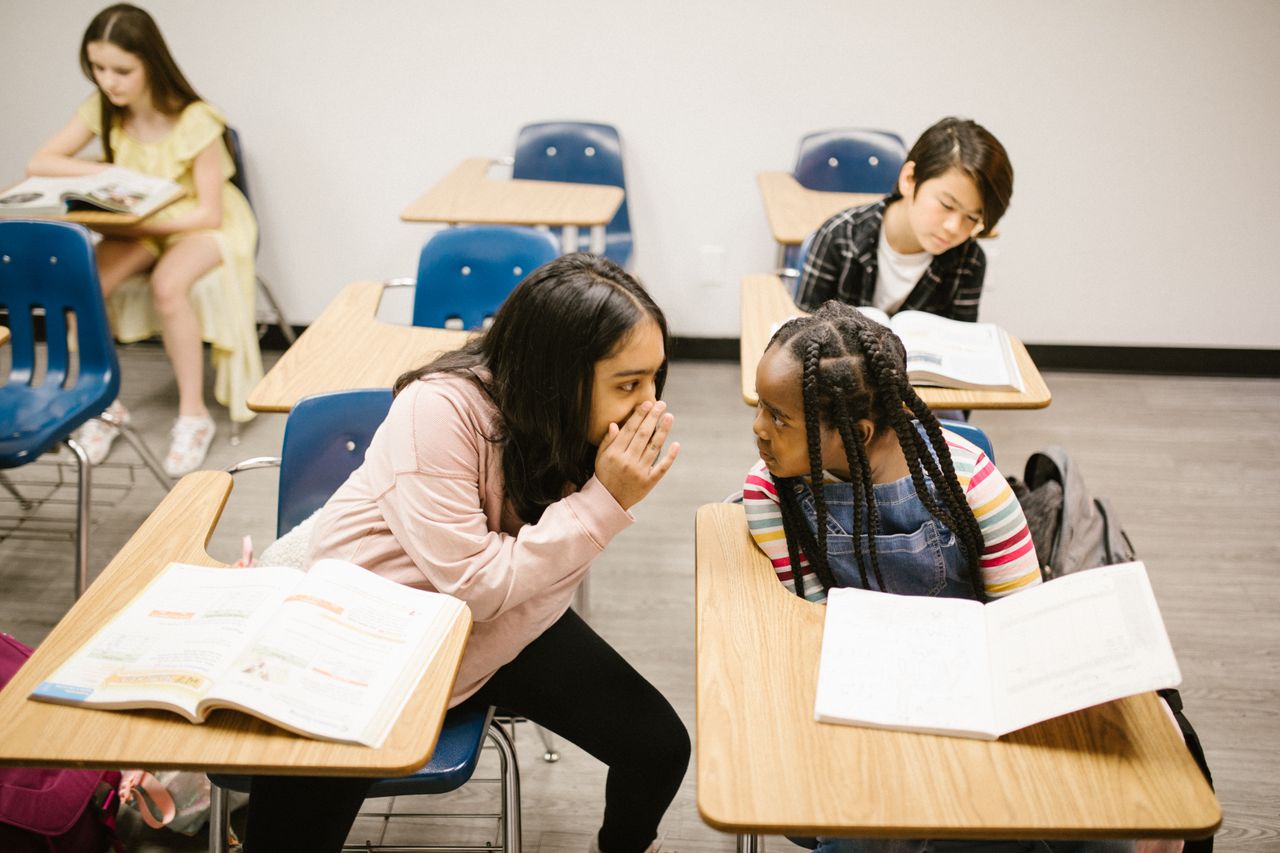 Three students in a classroom, two whispering, focusing on friendship and learning.