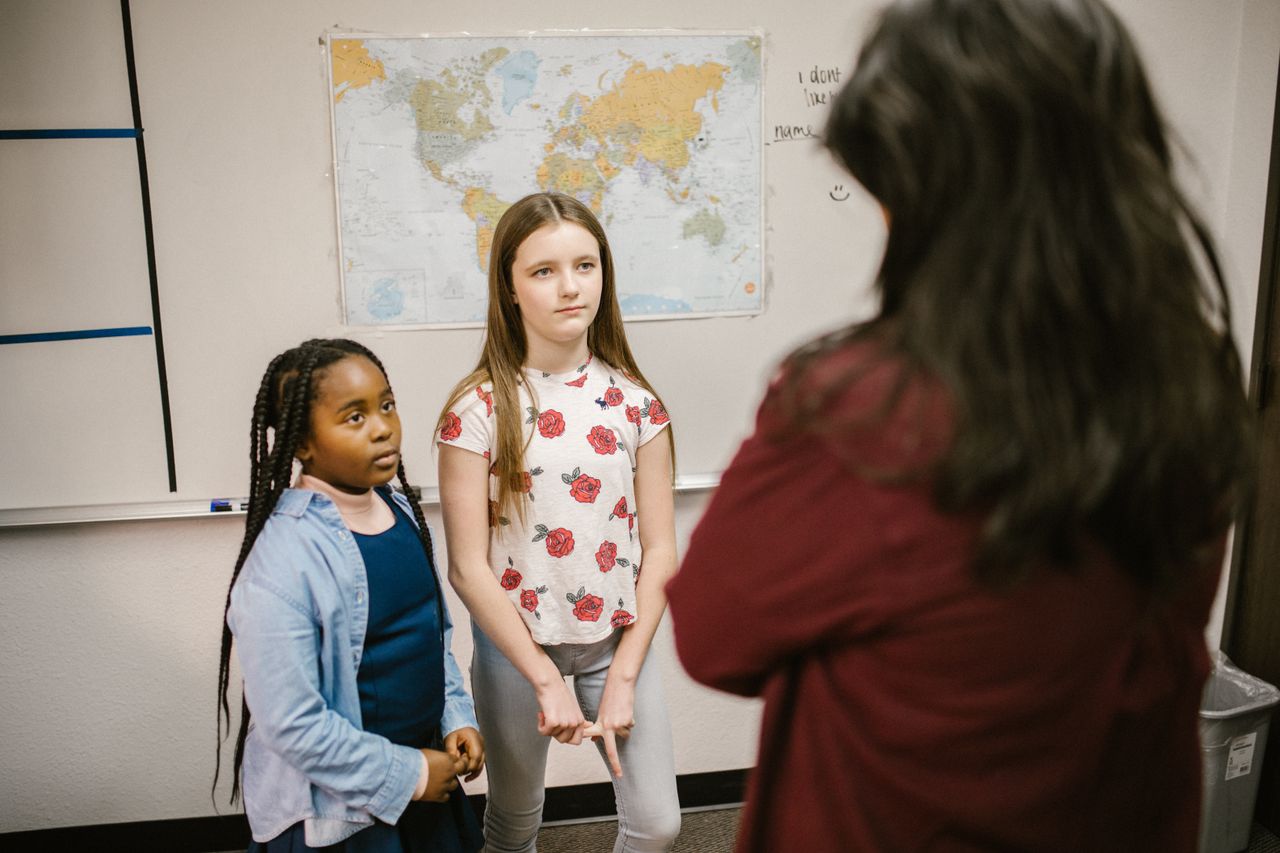 Two students stand in front of a teacher discussing bullying in a classroom setting.
